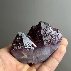 May include: A raw amethyst crystal cluster featuring two prominent, dark purple, pointed terminations with faceted surfaces. The base of the mineral specimen is opaque and lighter in colour, transitioning into deep, translucent violet peaks.