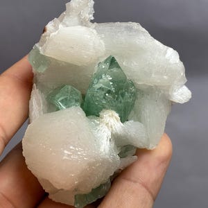 Könnte beinhalten: Ein Mineralexemplar mit durchscheinenden, blassgrünen, spitzen Kristallen, die in eine Ansammlung von undurchsichtigen, weißen, blockartigen und faserigen Kristallformationen eingebettet sind. Die Struktur zeigt einen Kontrast zwischen den grünen Spitzen.