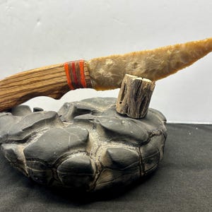 May include: A handcrafted knife with a wooden handle and a light-colored stone blade. The handle is wrapped with red and orange twine. The knife rests on a dark gray, textured stone base, and is supported by a small wooden piece.