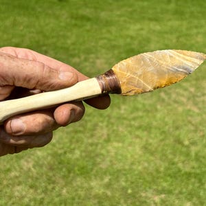 May include: A knife with a light tan, smooth bone handle and a sharp, pointed blade made of translucent, multi-colored flint stone. The blade is attached to the handle with dark brown cord wrapping, creating a distinct transition between the two parts.