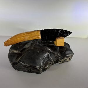 May include: A black obsidian knife with a light brown wooden handle, resting on a small wooden block support. The knife is positioned atop a large, textured, dark gray stone base with visible cracks and irregular surface patterns.