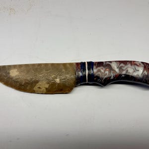May include: A handcrafted knife with a patterned blade and a handle featuring a swirl of red, white, and black. The blade has a textured surface. The handle has a dark blue and silver accent.