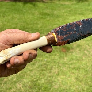 May include: A knife featuring a sharp, dark brown and black obsidian blade with a serrated edge. The handle is made of a smooth, light-colored bone material, securely attached to the blade with brown cord wrapping at the base of the blade.