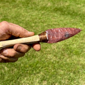 May include: A knife with a sharp, reddish-brown stone blade featuring a flaked texture. The handle is made of a light-colored, smooth bone, and the junction between the blade and the handle is wrapped with thin, dark brown cordage material.