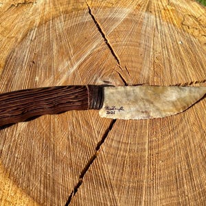 May include: A handcrafted knife with a dark wood handle and a light-colored, stone blade. The handle has a textured, striped pattern. The blade is signed and dated "2021". The knife is displayed on a wooden surface.