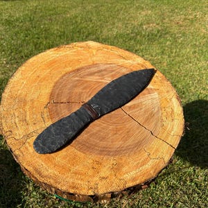 May include: A black, hand-crafted knife with a textured blade and wrapped handle rests on a cross-section of a tree trunk. The knife is positioned horizontally, showcasing its dark color and detailed craftsmanship. The background is green grass.