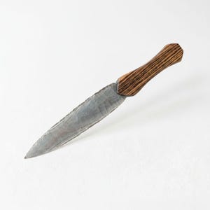 May include: A handcrafted knife with a gray, stone-like blade and a wooden handle. The handle features a striped pattern in shades of brown and tan. The blade is approximately 7 inches long, with a sharp, pointed tip.