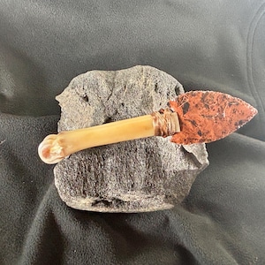 May include: A primitive-style knife with a bone handle and a reddish-brown, pointed blade. The blade is attached to the handle with a wrapped material. The knife rests on a gray, textured stone, suggesting a handcrafted or historical artifact.