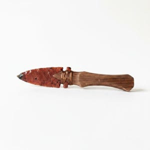 May include: A handcrafted arrowhead knife with a reddish-brown stone blade and a wooden handle. The handle is a dark brown color and is wrapped with a natural fiber. The knife is displayed on a white background.