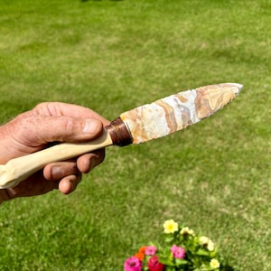 May include: A knife with a chipped, multi-colored stone blade featuring bands of white, tan, and brown. The handle is a smooth, light-colored material, connected to the blade by a dark, wrapped band that secures the two components together.