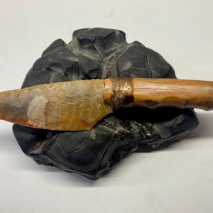 May include: A handcrafted flint knife with a wooden handle, resting on a dark, textured stone. The blade is a light brown color with darker marbling, and the handle is a lighter wood, bound with a dark fiber.