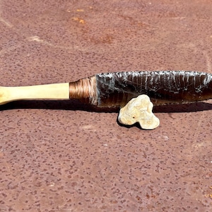 May include: A knife featuring a dark, serrated obsidian blade attached to a smooth, light-colored bone handle. The connection point is wrapped in thin, dark brown cordage. A small, irregular, light-colored stone piece supports the blade.