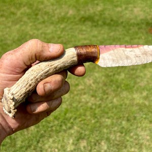 May include: A knife featuring a curved, textured antler handle and a sharp, light-colored stone blade with a pinkish hue along the top edge. The blade is securely attached to the handle with dark brown cord wrapping and a visible amber-colored adhesive.