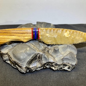 May include: A handcrafted knife with a wooden handle and a stone blade. The handle features a zebra wood pattern and is accented with red, blue, and black bands. The blade is a light yellow color and has a serrated edge. The knife rests on a dark gray stone.