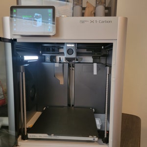 May include: A white and gray 3D printer with a black print bed and a clear plastic cover. The printer has a touchscreen display and a small camera on the top. The printer is labeled "Bamboo Lab X1-Carbon".