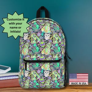 May include: A black backpack with a colorful pattern of aliens, cats, and other whimsical characters. The backpack has a black zipper and a top handle. The text "Customize it with your name or initials!" is in a green starburst. Made in USA.