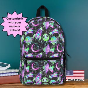 May include: Black backpack with a colorful space-themed pattern featuring rockets, planets, aliens, and stars. The backpack has a front pocket and a top handle. Text on the image says "Customize it with your name or initials!" and "Made in USA".