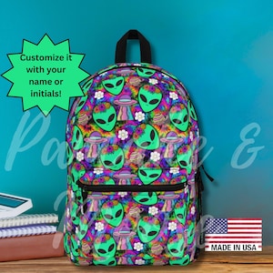 May include: A backpack with a vibrant pattern of green alien faces, UFOs, and flowers on a tie-dye background. The backpack has a black zipper and handle. Text on the image says "Customize it with your name or initials!" and "Made in USA".