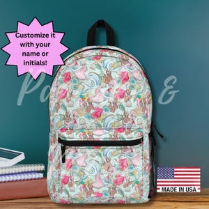 以下が含まれることがあります： ピンク、青、緑色の人魚模様のバックパック。バックパックには黒いジッパーと黒いハンドルが付いています。「Customize it with your name or initials!」というテキストはピンク色のスターバーストにあります。「Made in USA」という言葉が表示されています。