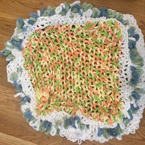 May include: Handmade knitted blanket with a square center and a ruffled edge. The center features a mix of orange, green, and yellow yarn. The border is white with blue and green accents. A cozy, decorative item.