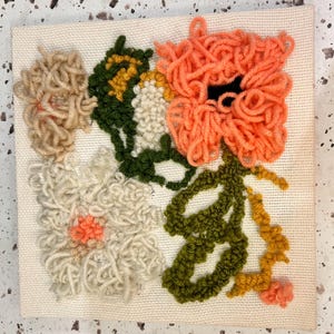 May include: A close-up of a handmade wall hanging featuring a floral design with orange, white, green, and yellow yarn. The flowers are made with a fluffy, looped technique.