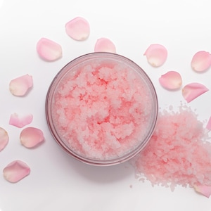 May include: A glass jar filled with pink sugar scrub, surrounded by pink rose petals on a white background.