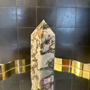 Op de afbeelding: Een hoge, rechthoekige, gepolijste stenen obelisk met een puntige top. De steen heeft een mix van witte, bruine en groene patronen. De obelisk wordt op een reflecterend oppervlak weergegeven. Twee prijskaartjes zijn zichtbaar.