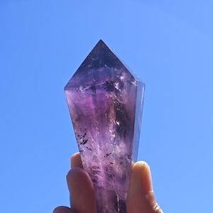 Puede incluir: Una gran punta de cristal de amatista púrpura con una forma facetada clara. El cristal se sostiene en una mano contra un cielo azul brillante.