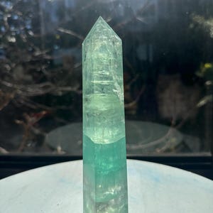 Puede incluir: Una torre de cristal de fluorita hexagonal con una punta afilada. La piedra presenta capas translúcidas de tonos verde menta pálido y verde esmeralda más intenso, con fracturas internas visibles e inclusiones naturales en la estructura.