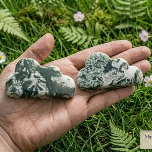 May include: Two cloud-shaped carvings made of moss agate, featuring a smooth, polished surface with distinct dark green and white marbled patterns. The stones are displayed together, showcasing their organic, swirling inclusions and rounded edges.