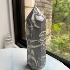 May include: A grey and white marble obelisk with a pointed top. The stone has a swirling pattern of grey and white. The obelisk is standing on a white surface near a window.