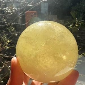 May include: A translucent, yellow crystal ball is held in a hand. The sphere is smooth and round, reflecting light. The background shows a window with greenery and a blurred reflection.