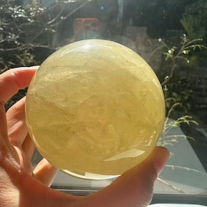 May include: A large, translucent yellow crystal sphere, held in a hand. The sphere has a smooth, polished surface and appears to be made of a natural stone. The light shines through the sphere, creating a glowing effect. The background is blurred.