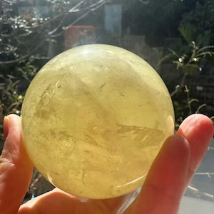 May include: A large, translucent yellow crystal sphere, held in a hand. The sphere is smooth and appears to have internal patterns. The background is blurred, with hints of greenery and sunlight. This crystal ball is a decorative item.