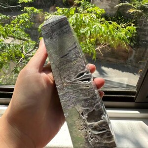 May include: A rectangular, multi-toned stone obelisk with a pointed top. The stone features a mix of grey, green, and white hues with natural patterns and textures. The obelisk is held in a hand, with a window and greenery in the background.