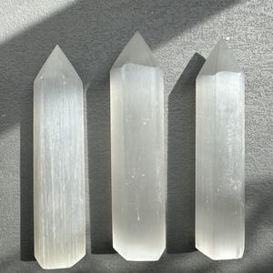 May include: Three white selenite crystal wands of varying sizes, arranged horizontally on a grey surface.