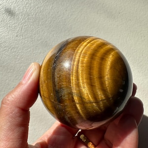 May include: A polished tiger's eye sphere, displaying bands of golden brown and dark brown colours. The gemstone ball is held in a hand, with sunlight highlighting its natural patterns and textures. A decorative item.