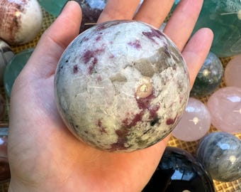 May include: A hand holds a large, round, white and pink crystal sphere. Other crystal spheres in shades of green, pink, and black are in the background, along with a woven basket. The sphere has a marbled appearance with pink and gray inclusions.