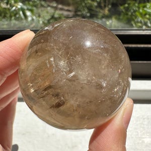 May include: A clear quartz crystal sphere with a smoky brown hue. The sphere is smooth and polished, with visible internal inclusions.