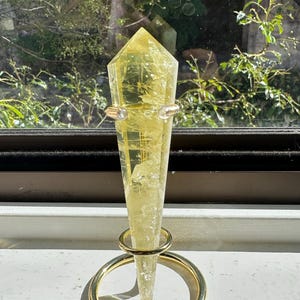 Könnte beinhalten: Ein facettierter, gelber Kristallpunkt wird in einem goldfarbenen Metallständer präsentiert. Der Kristall ist ein helles, durchscheinendes Gelb. Der Ständer hat einen kreisförmigen Sockel und einen Ring, der den Kristall nahe der Spitze hält. Der Hintergrund zeigt ein Fenster mit grünem Laub.