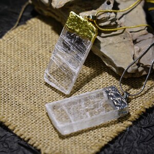 May include: Two rectangular crystal pendants with metal accents, one gold and one silver, on delicate chains. The pendants rest on a burlap surface, with a dark background. The crystals have a clear, textured appearance.