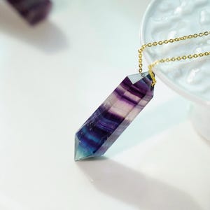 Könnte beinhalten: Ein violetter, blauer und weißer Fluorit-Kristall-Anhänger an einer goldenen Kette. Der Kristall ist facettiert und hat eine spitze Spitze.