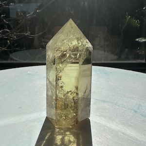 Könnte beinhalten: Eine durchscheinende, hexagonale Citrin-Kristallspitze mit einer spitzen Oberseite. Der Stein weist einen blassgelben Farbton und interne Risse auf, die ein texturiertes, kristallines Erscheinungsbild im gesamten Mineralexemplar erzeugen.