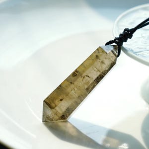 May include: A clear quartz crystal point pendant on a black cord necklace. The crystal is a light brown color and has a pointed tip. The pendant is hanging from a white surface.