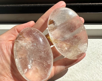 May include: Two clear quartz crystal palm stones with a light brown inclusion. The stones are smooth and polished, and they are held in a hand.