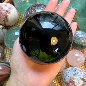 May include: A black obsidian sphere, polished to a high shine, is held in a hand. The sphere reflects light and shows a slight green hue. The sphere is surrounded by other polished spheres of various colours, including green, pink, and white.