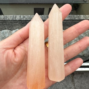 May include: Two pink and white crystal points, also known as selenite wands, are held in a hand. The wands are smooth and have a slight shimmer.