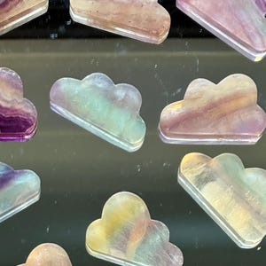 May include: A collection of nine iridescent rainbow fluorite cloud shaped crystal carvings. The crystals are translucent and have a variety of colors, including purple, pink, green, and yellow.