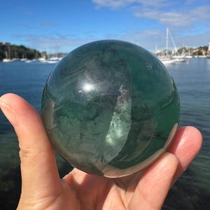 May include: A large, green fluorite sphere with white and gray striations. The sphere is being held in a hand.