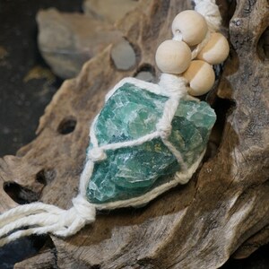 May include: A teal-colored fluorite crystal pendant, wrapped in white macrame cord, is adorned with wooden beads. The crystal is displayed on a piece of weathered driftwood, creating a natural and earthy aesthetic. The pendant is designed to be worn as a necklace.